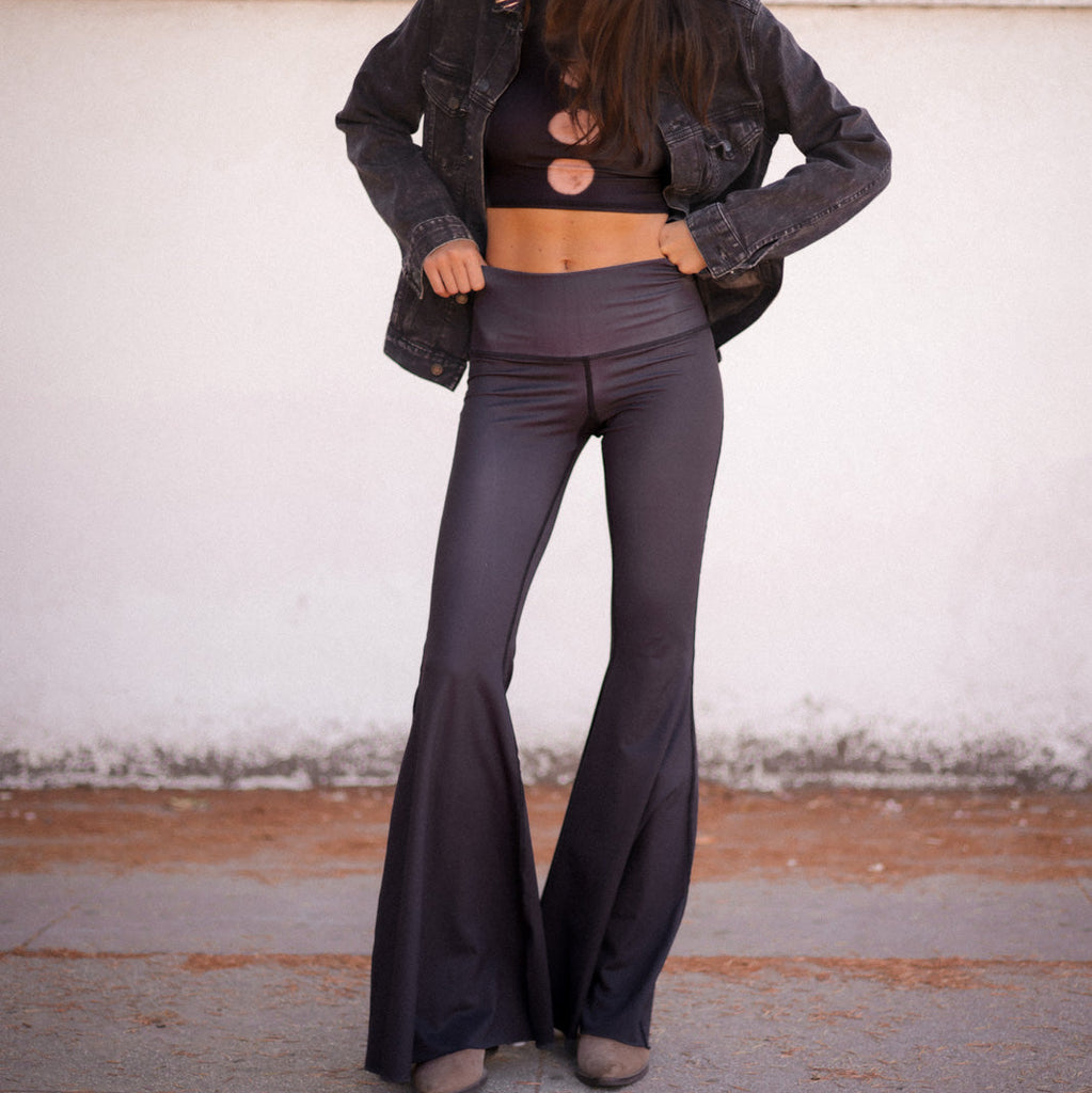 Person wearing a dark jacket, crop top, and flared pants against a plain background